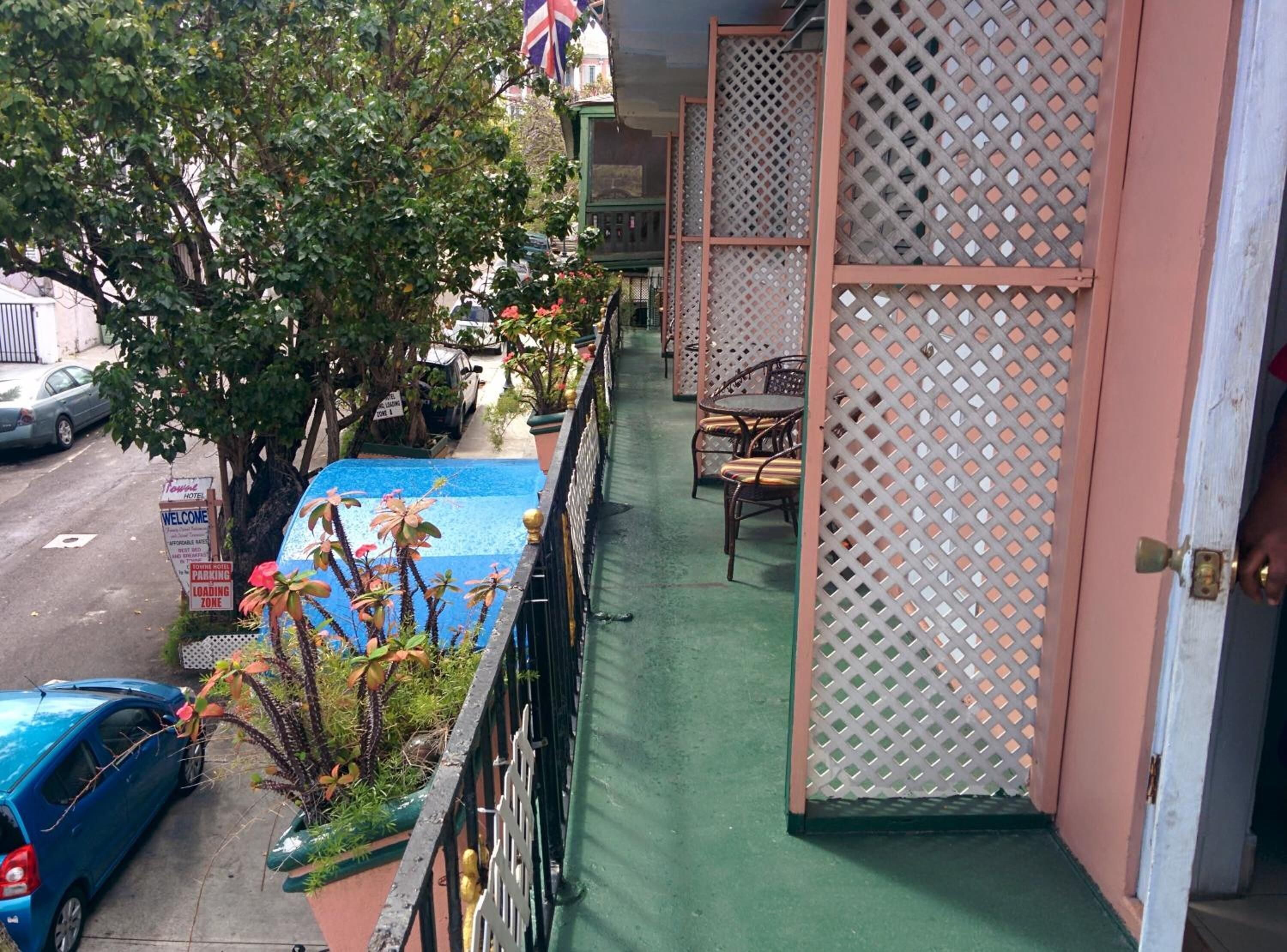 Un balcón con piso verde, una mesa con sillas y vista a una calle con autos y un edificio con una bandera.