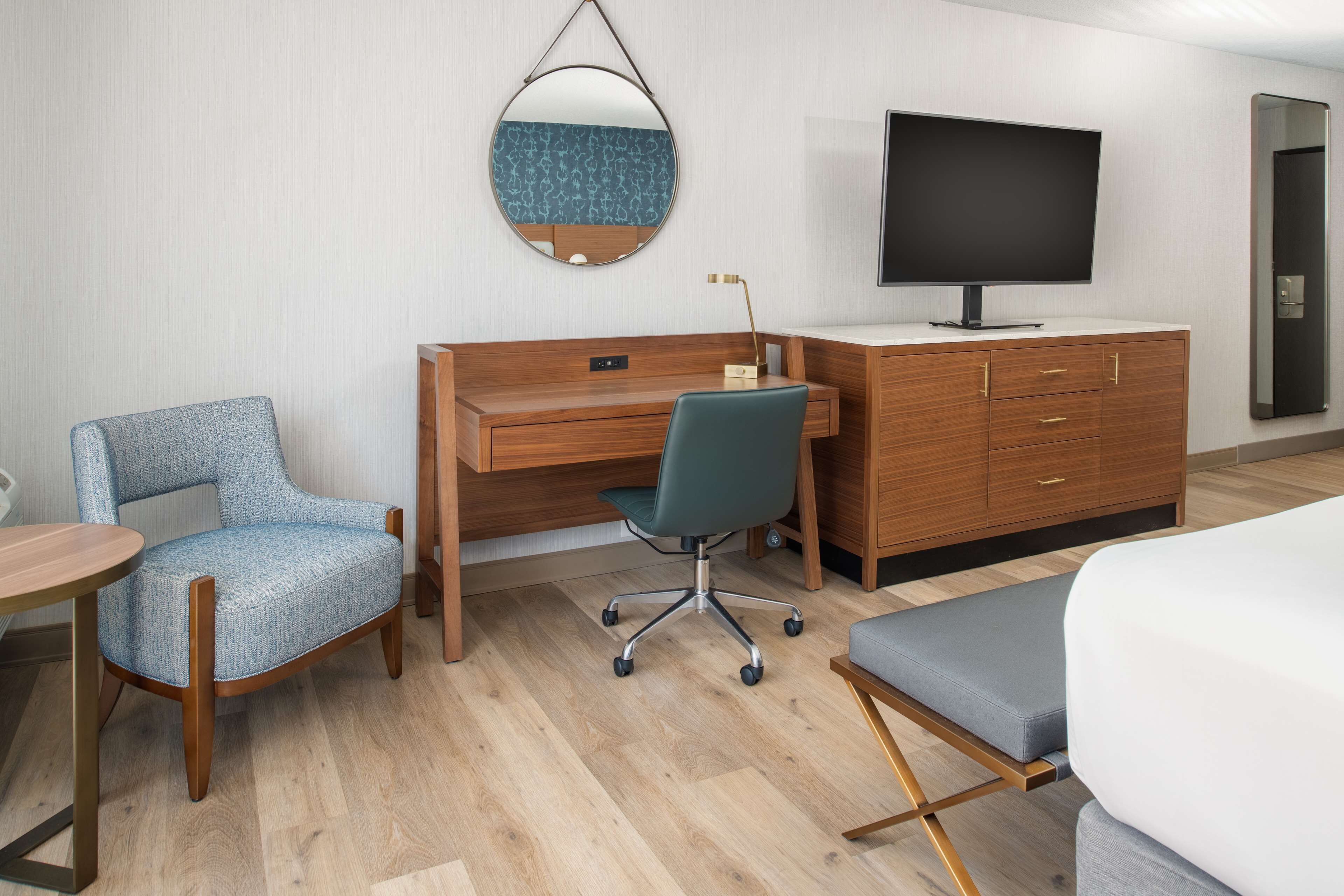 A hotel room with a wooden desk, a blue chair, a television, and a bed.