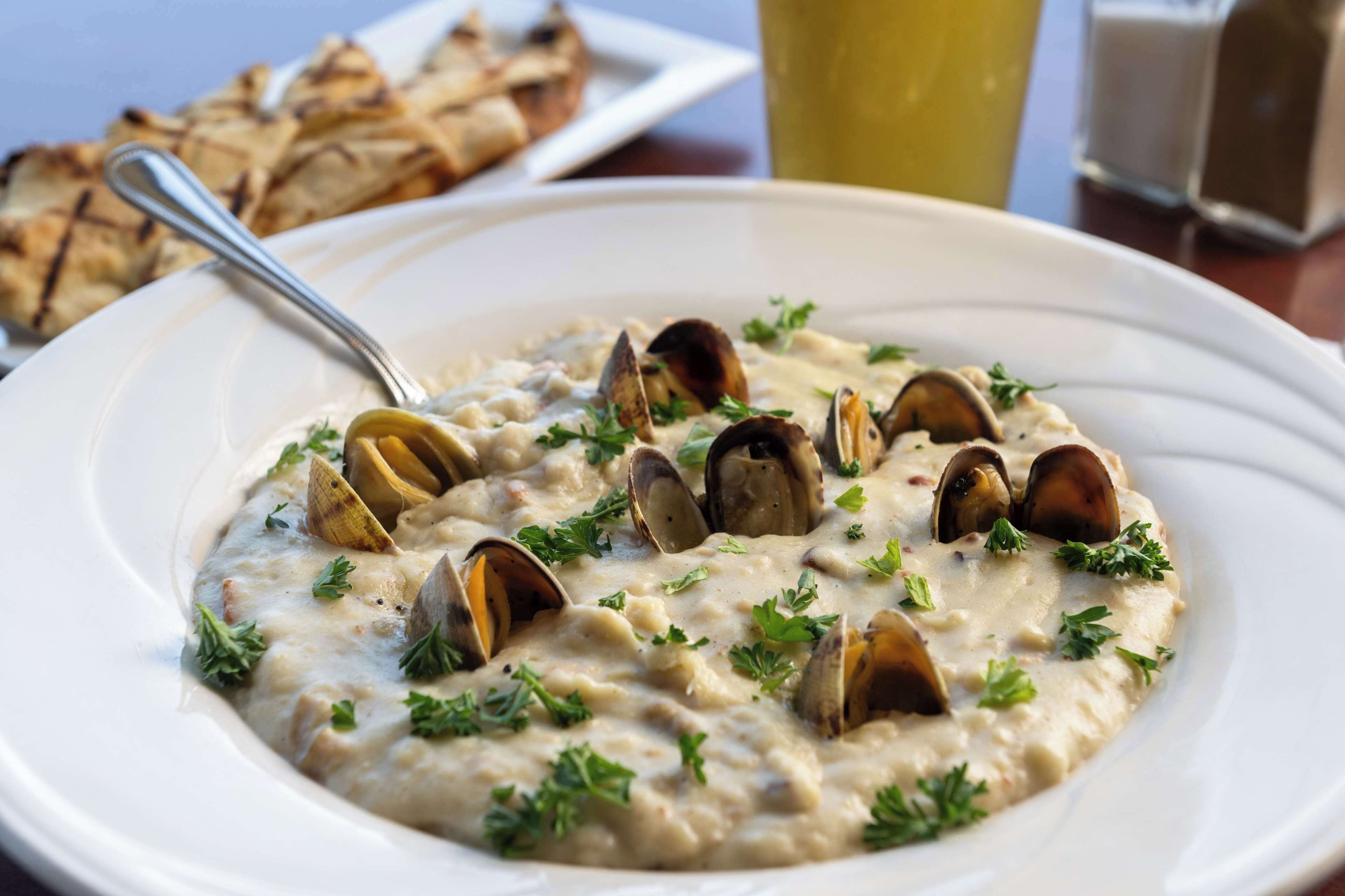 A white plate with a creamy seafood dish garnished with parsley, accompanied by grilled bread and a glass of beverage.