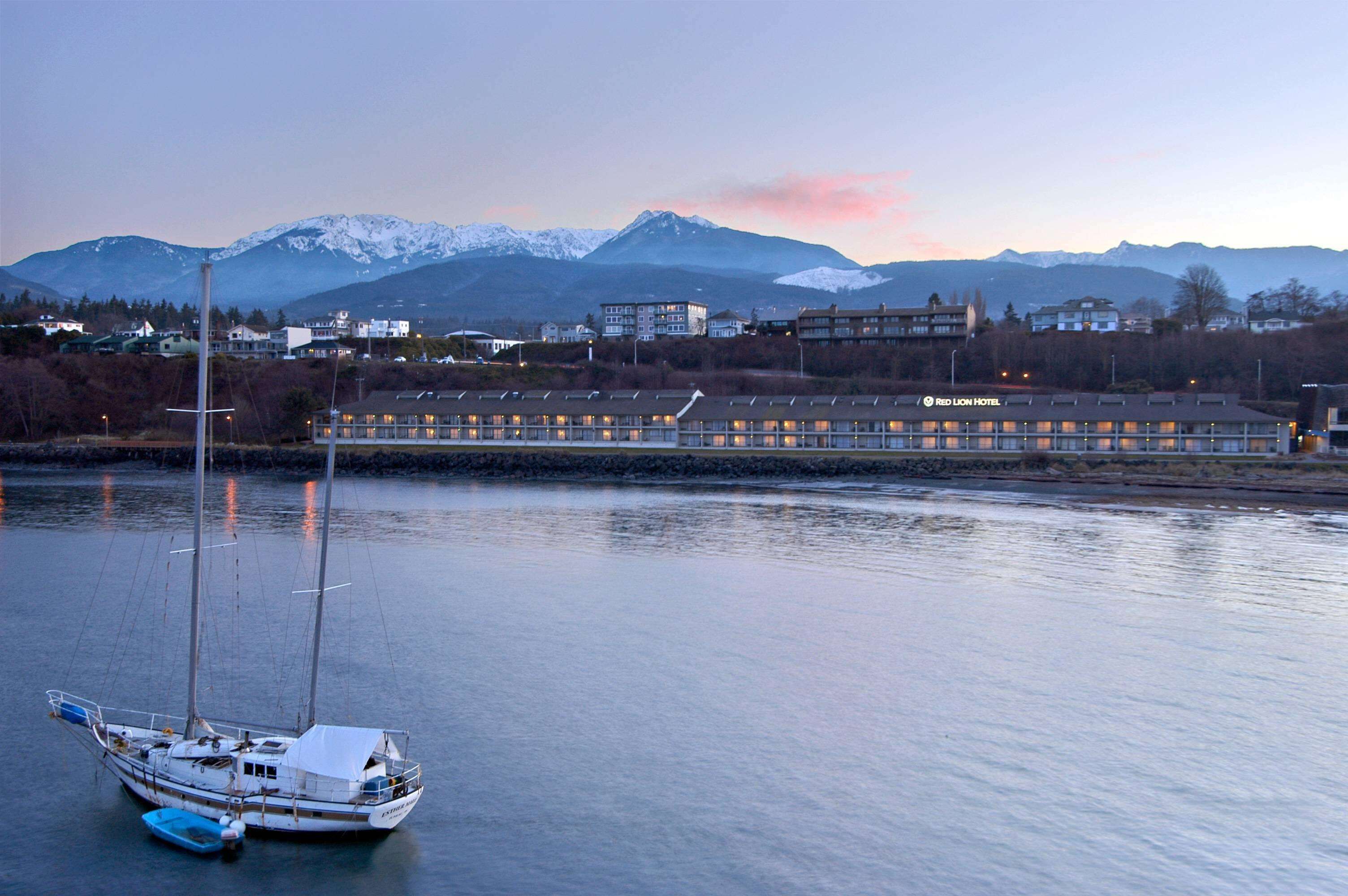 A waterfront hotel with a sailboat docked in the foreground, a mountain range in the background, and a clear sky.