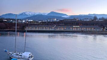 A waterfront hotel with a sailboat docked in the foreground, a mountain range in the background, and a clear sky.