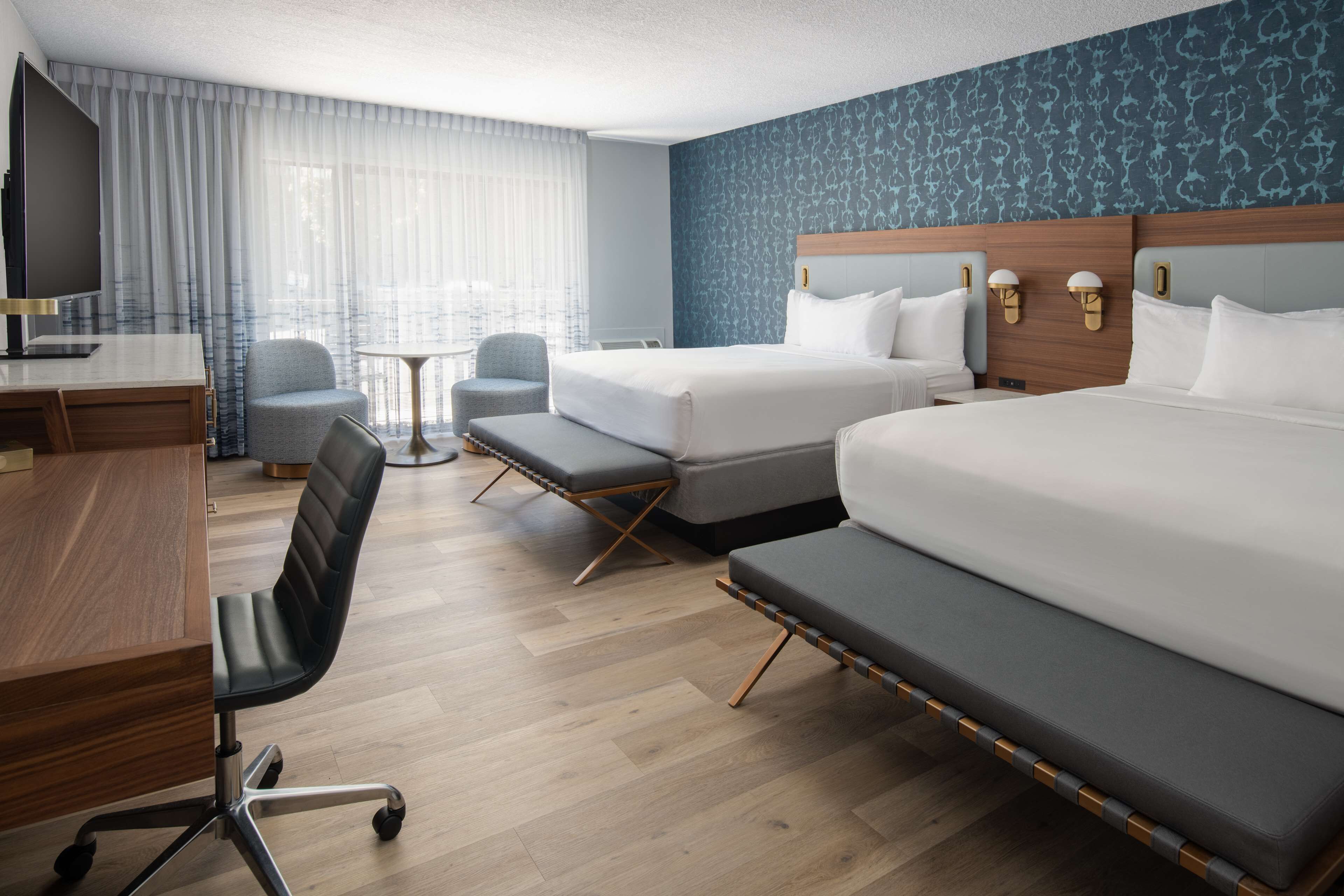 A hotel room with two beds, a desk, a chair, a small table, and a TV.