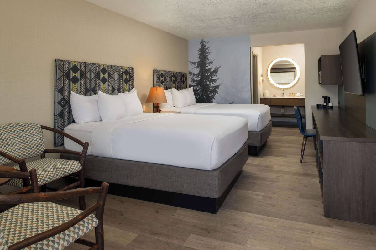 A hotel room with two beds, a desk, a chair, a TV, and a bathroom visible through an open door.