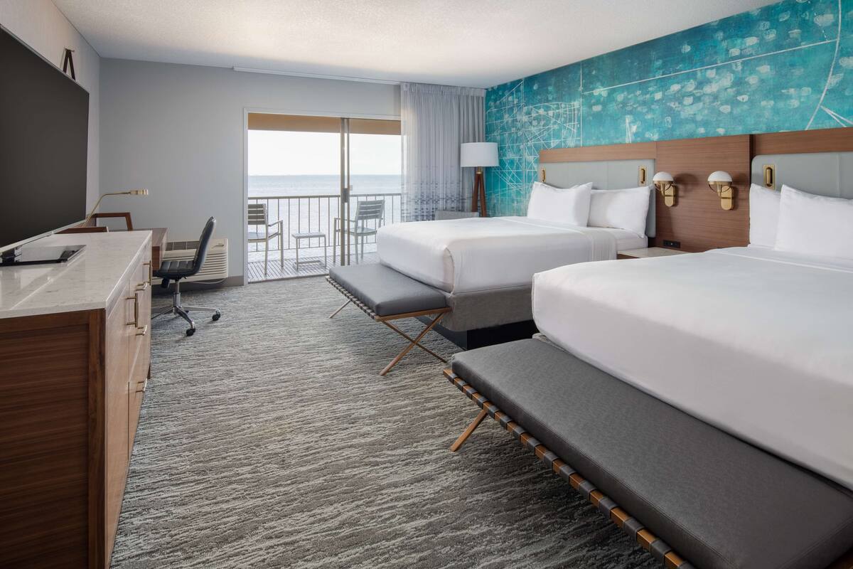 A hotel room with two beds, a desk with a chair, a large window with a view of the ocean, and a blue textured wall.