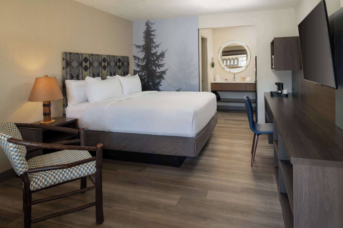 A hotel room with a bed, a desk, a chair, a lamp, a mirror, and a TV.