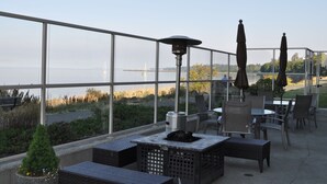 An outdoor patio with tables and chairs, a fire pit, and a view of the water and distant buildings.