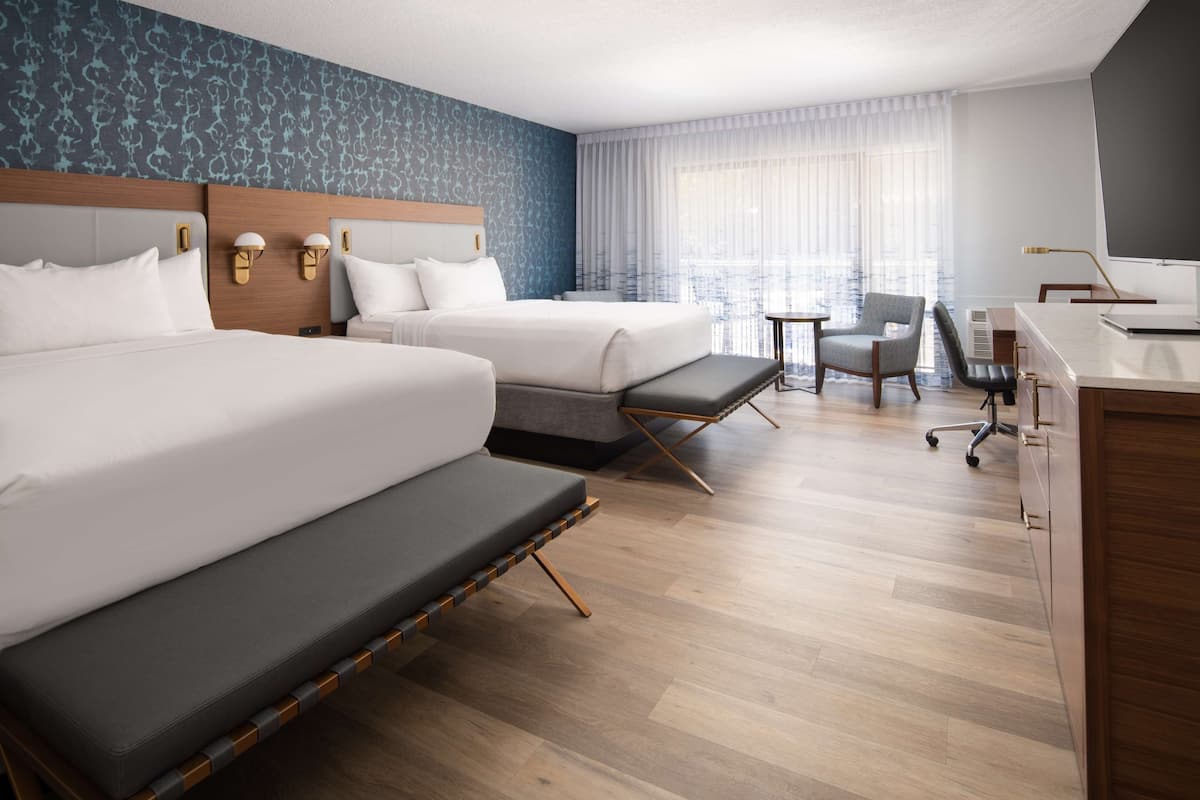 A hotel room with two beds, a desk, a chair, and a large window with curtains.