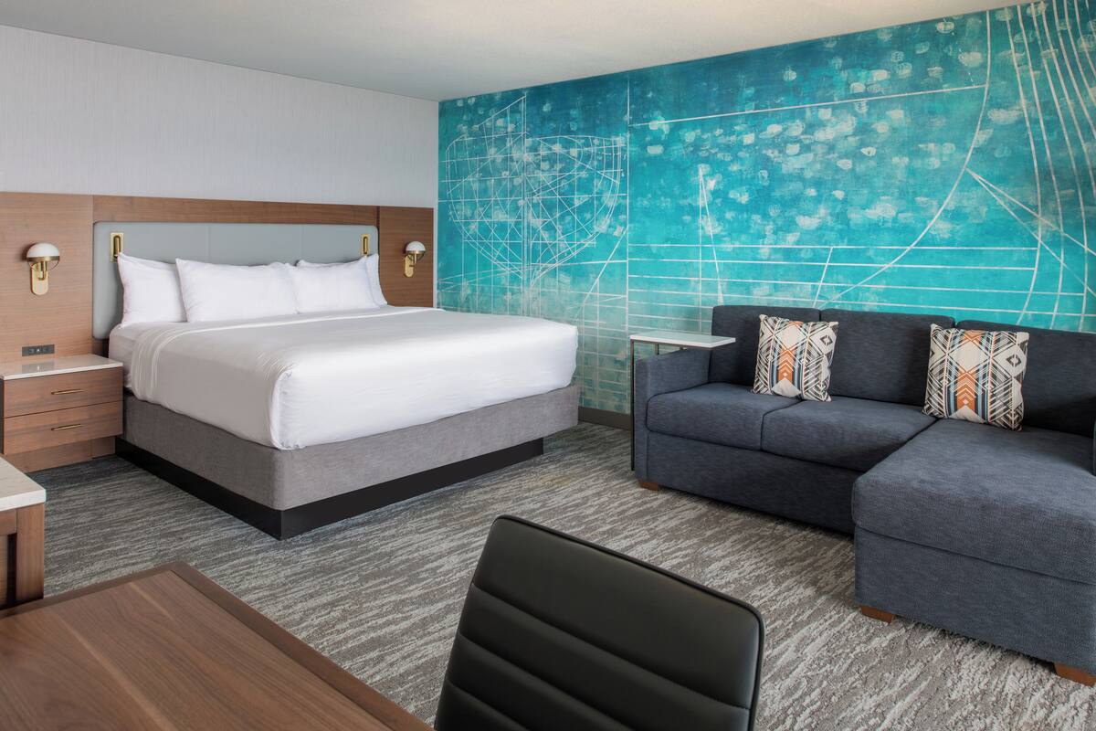 A hotel room with a large bed, a gray sofa, a small table, and a wall with a geometric design.
