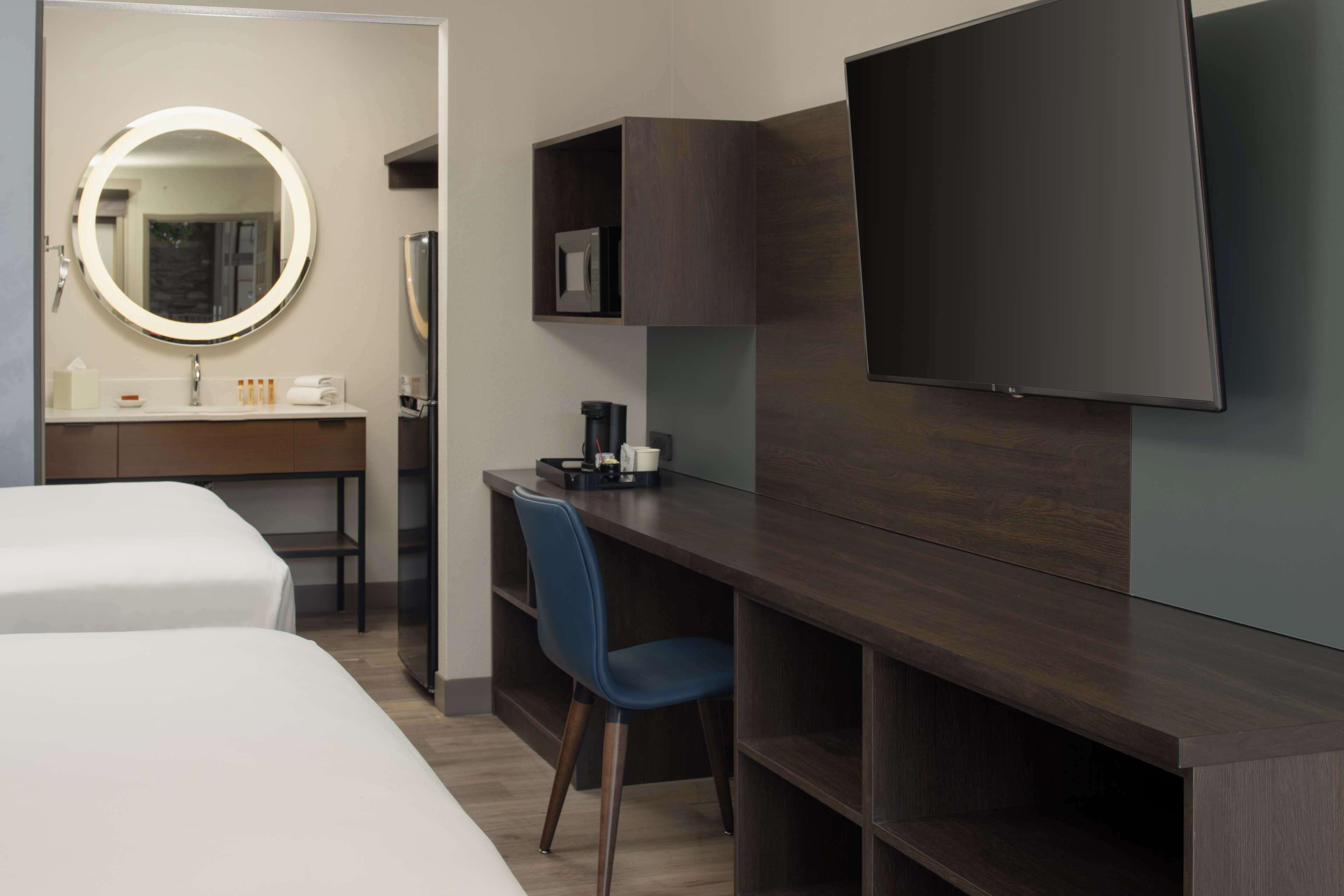 A hotel room with a bed, a desk with a chair, a TV, a mirror, and a small shelf with toiletries.