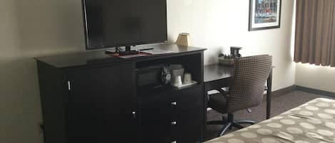 Ett hotellrum med en säng, ett skrivbord med en tv, en stol och en kaffebryggare.