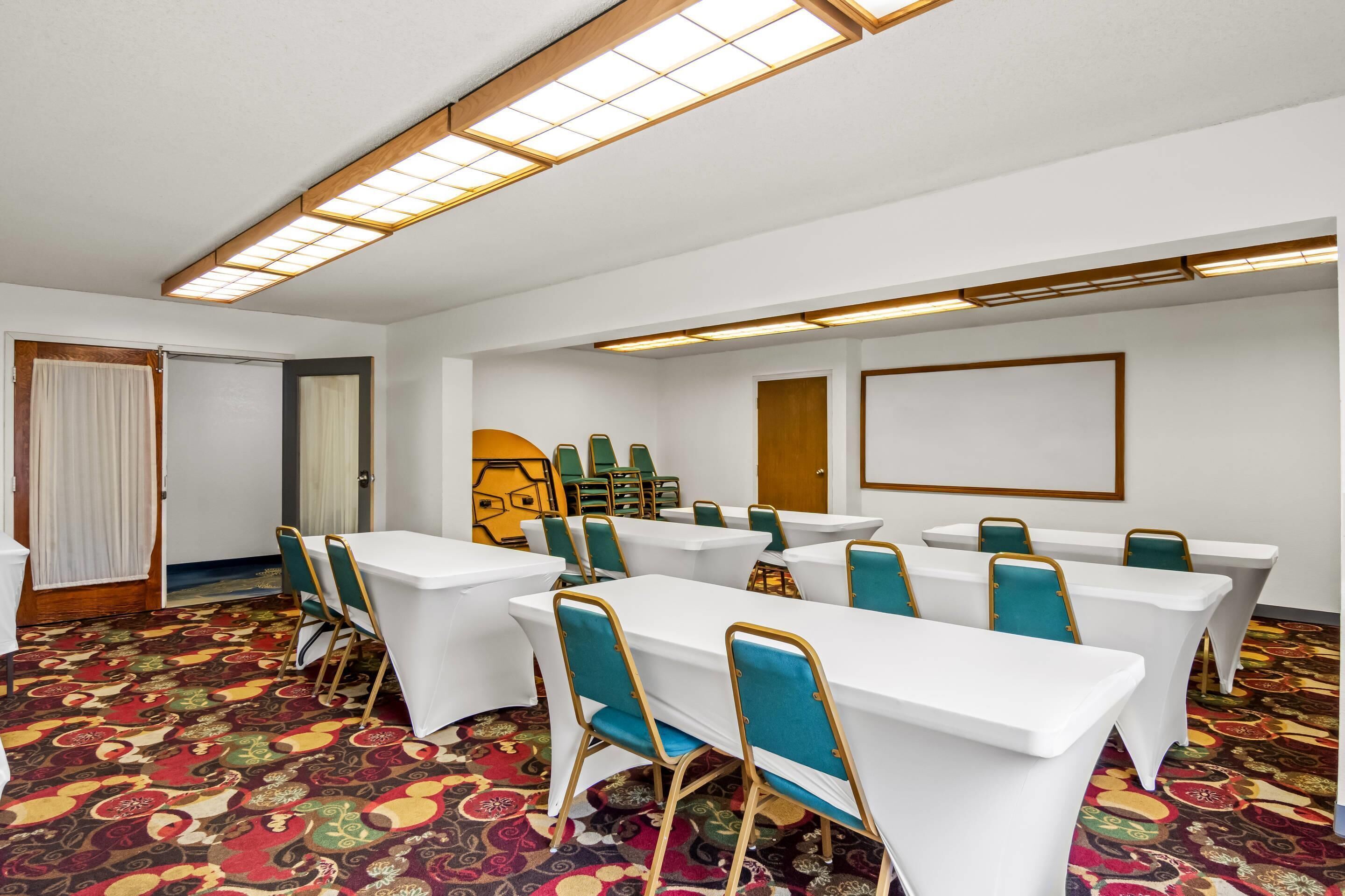 A conference room with white tables and teal chairs, patterned carpet, and a large screen on the wall.