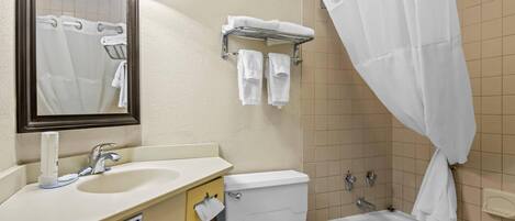 Baño con lavamanos, inodoro y bañera. Se ve un perchero con toallas y una cortina de ducha.