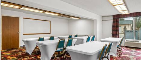 Une salle de conférence avec des tables blanches, des chaises de couleur turquoise, une moquette à motifs et une grande fenêtre avec des rideaux.