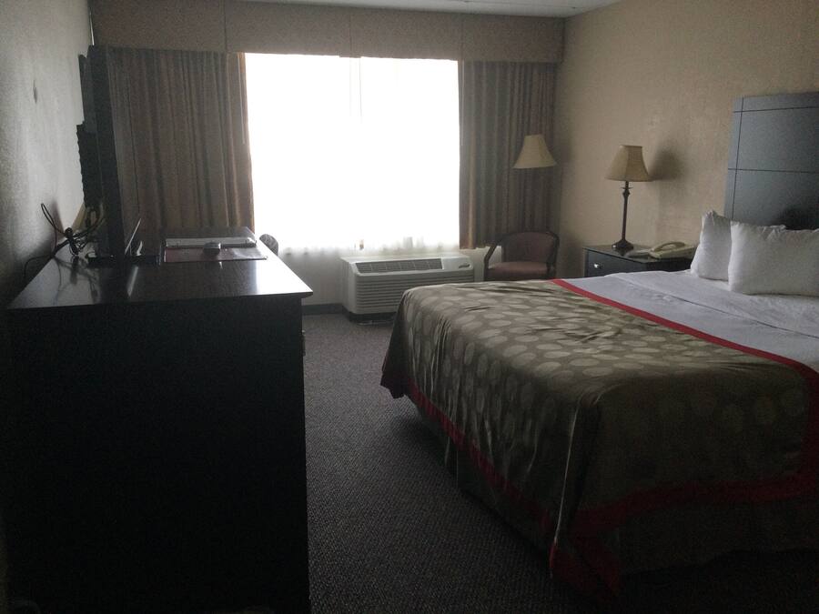 A hotel room with a bed, a desk, an air conditioner, a chair, and a lamp.