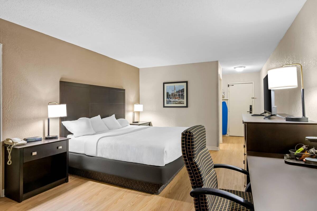 A hotel room with a bed, bedside tables, a desk, a chair, a desk lamp, a picture on the wall, and a door in the background.