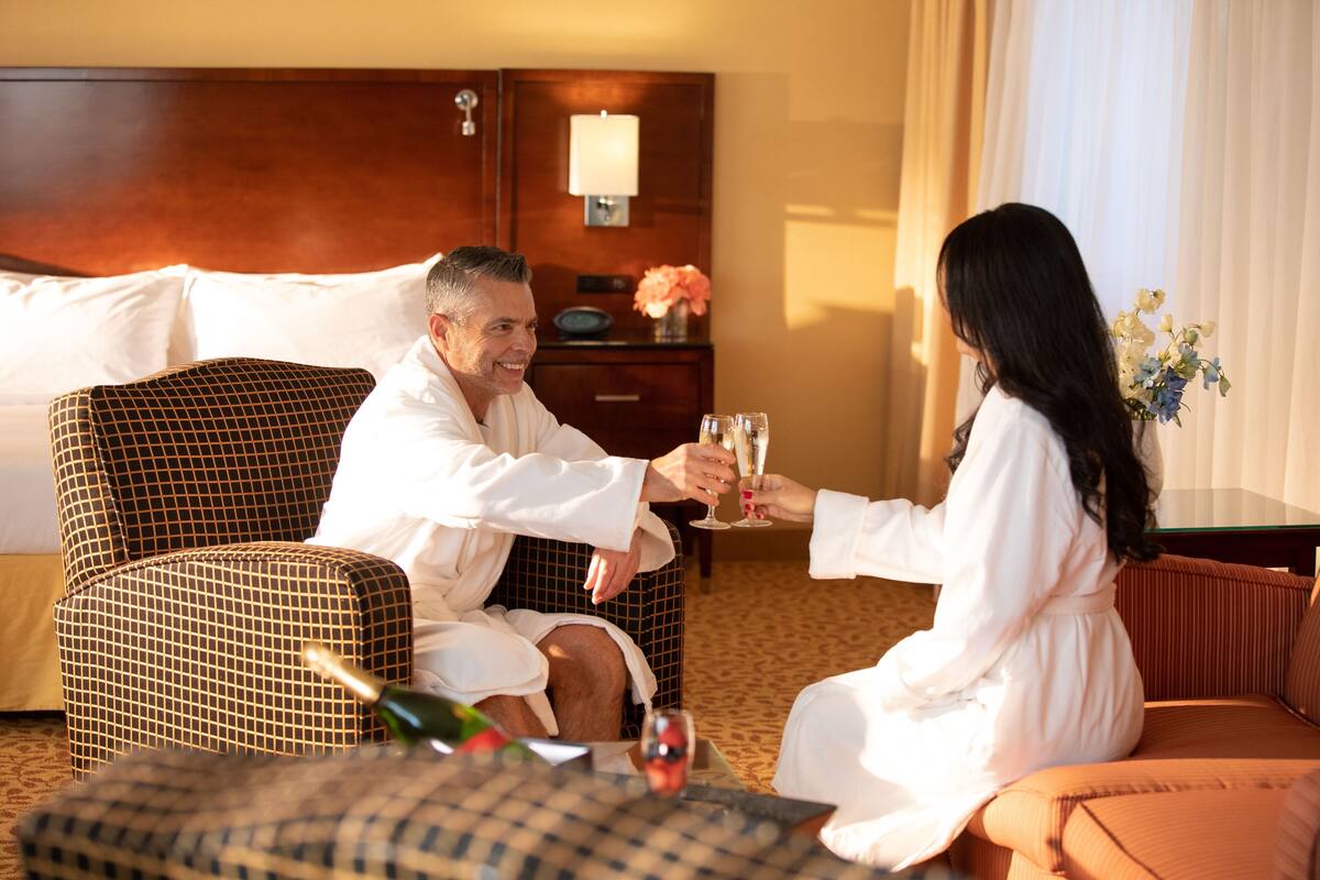 Una pareja en una habitación de hotel, sentada en un sofá, brindando con copas de champán.