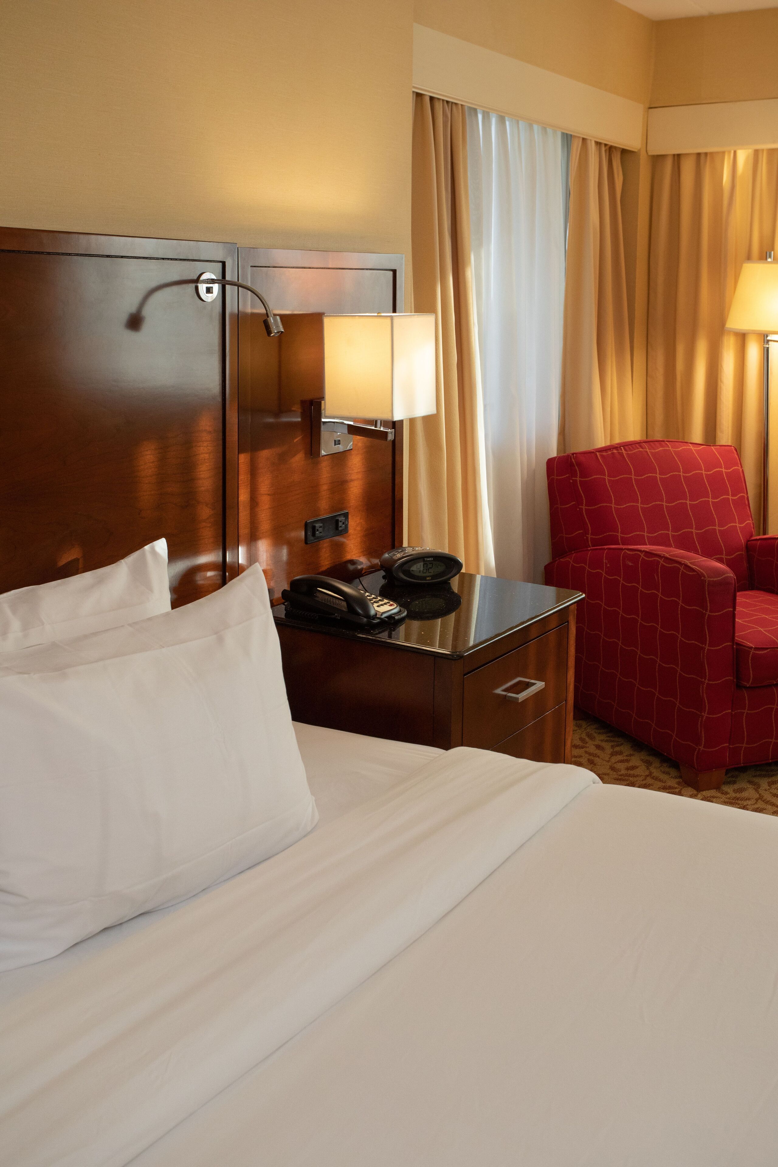 A hotel room with a bed, a nightstand with a lamp, a telephone, and a red chair.