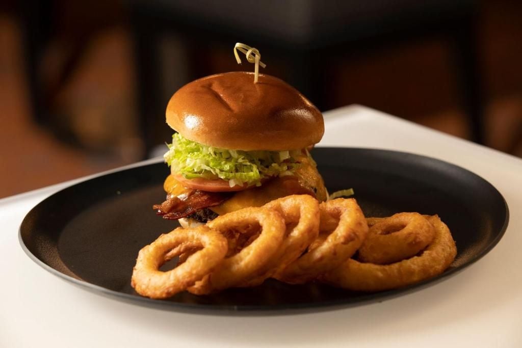 Un hamburger composé d’un pain, de laitue, de tomate et d’une tranche de bacon, servi avec des rondelles d’oignon dans une assiette.