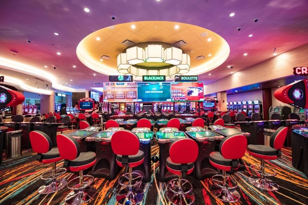 Un casino con múltiples mesas de blackjack y ruleta, sillas rojas y un candelabro central.