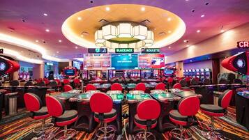 Un casino con múltiples mesas de blackjack y ruleta, sillas rojas y un candelabro central.