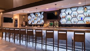 A bar area with a hexagonal shelving unit displaying bottles, a flat-screen TV, and bar stools. - Jake's 58 Casino Hotel - Adult Only (Islandia)