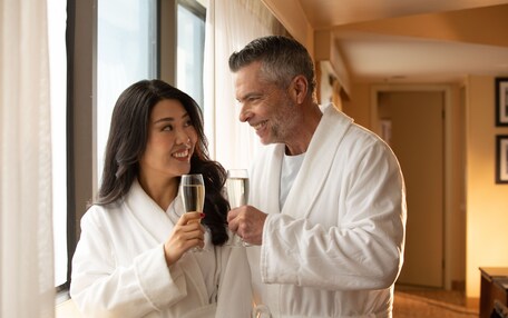 A couple in white bathrobes holding champagne glasses in a hotel room with a large window and a door in the background.