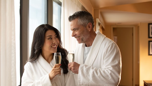 A couple in white bathrobes holding champagne glasses in a hotel room with a large window and a door in the background. - Jake's 58 Casino Hotel - Adult Only (Islandia)