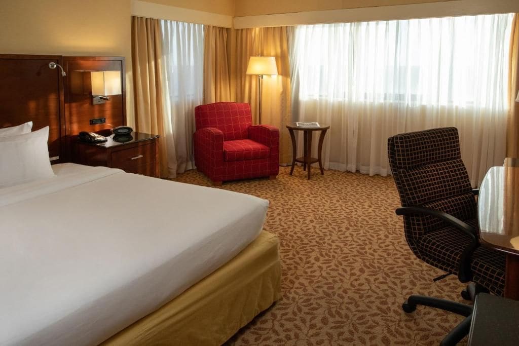 Een hotelkamer met een groot bed, een rode fauteuil, een zwarte bureaustoel, een klein tafeltje met een lamp en een nachtkastje met een telefoon.
