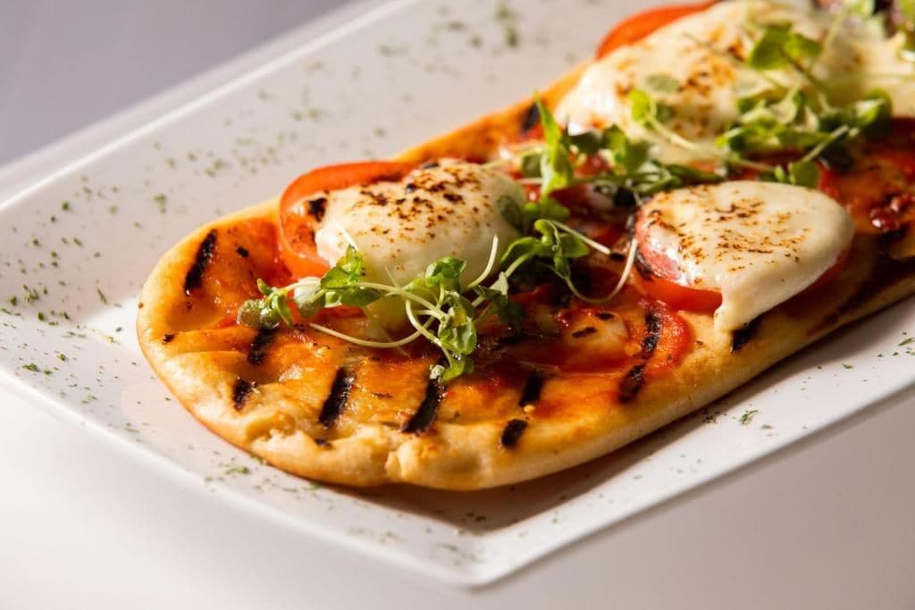 A rectangular plate with a grilled pizza topped with tomatoes, cheese, and greens.