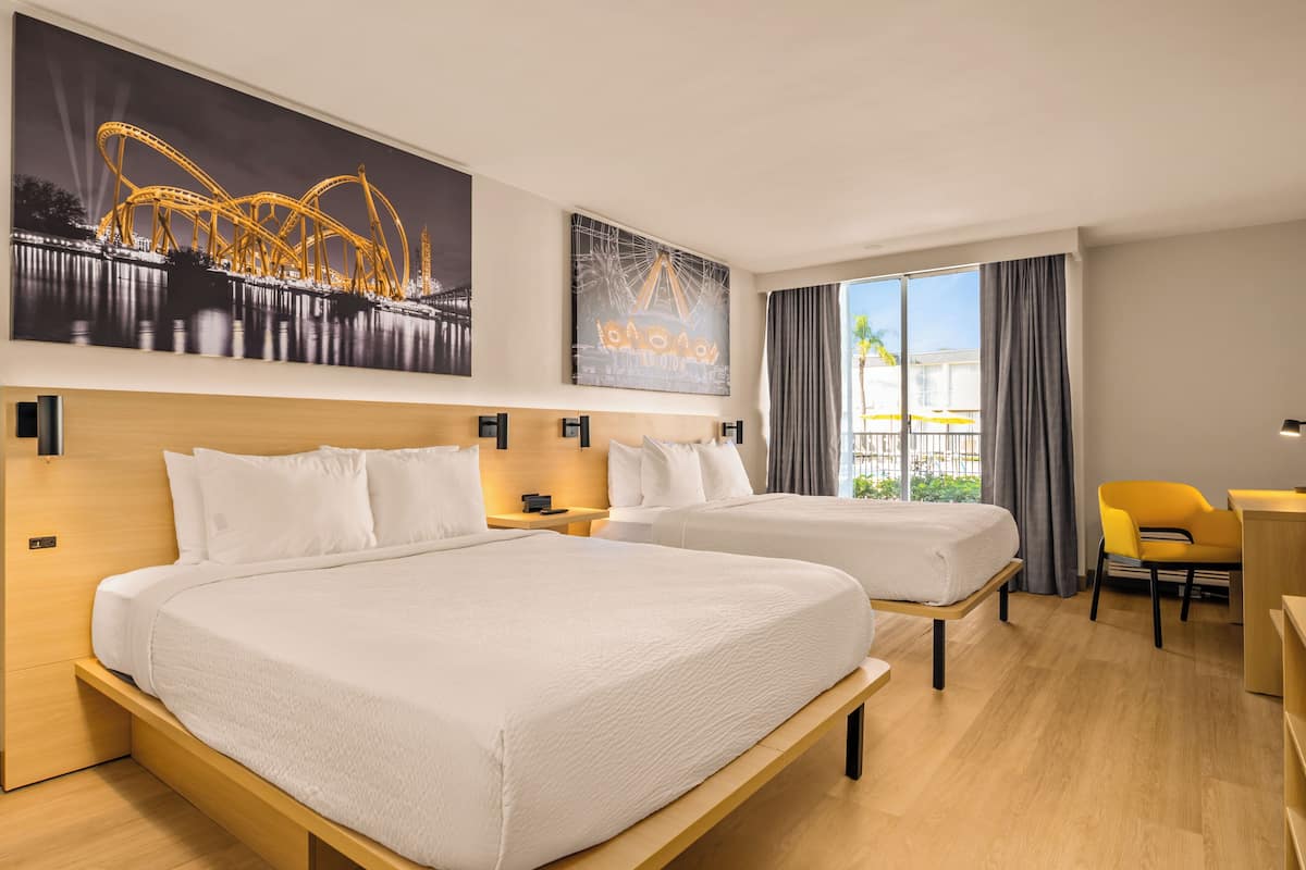 A hotel room with two beds, a desk, a chair, and large windows with a view of a building and palm trees.