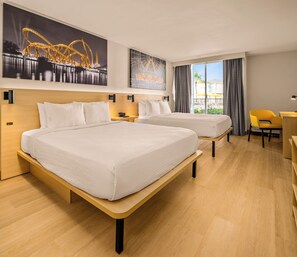 Quarto de hotel com duas camas, uma mesa, uma cadeira e janelas amplas com vista para um edifício e palmeiras.
