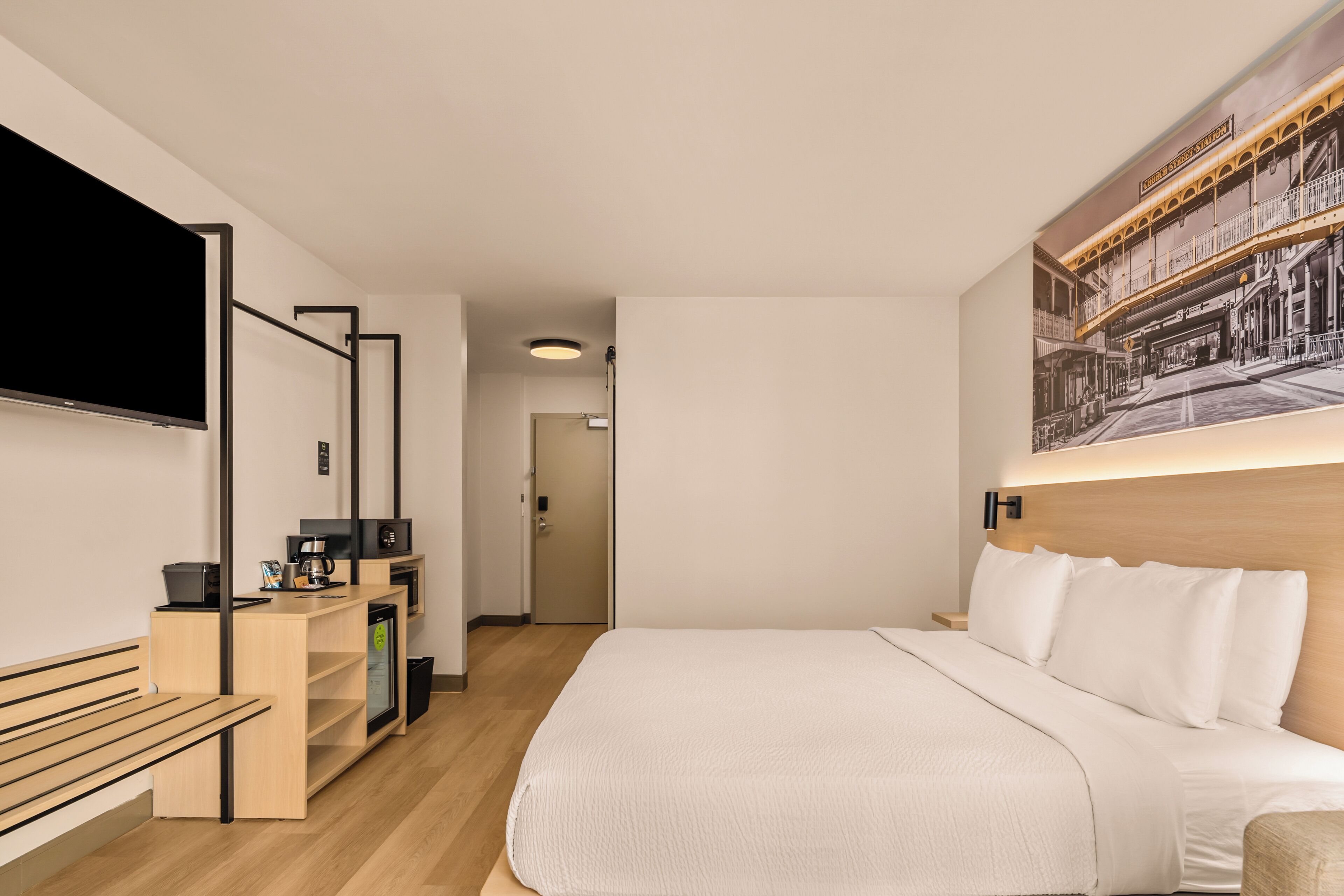 Una habitación de hotel moderna con una cama grande, un televisor de pantalla plana, un banco de madera, un pequeño escritorio con una cafetera y una obra de arte colgada en la pared.
