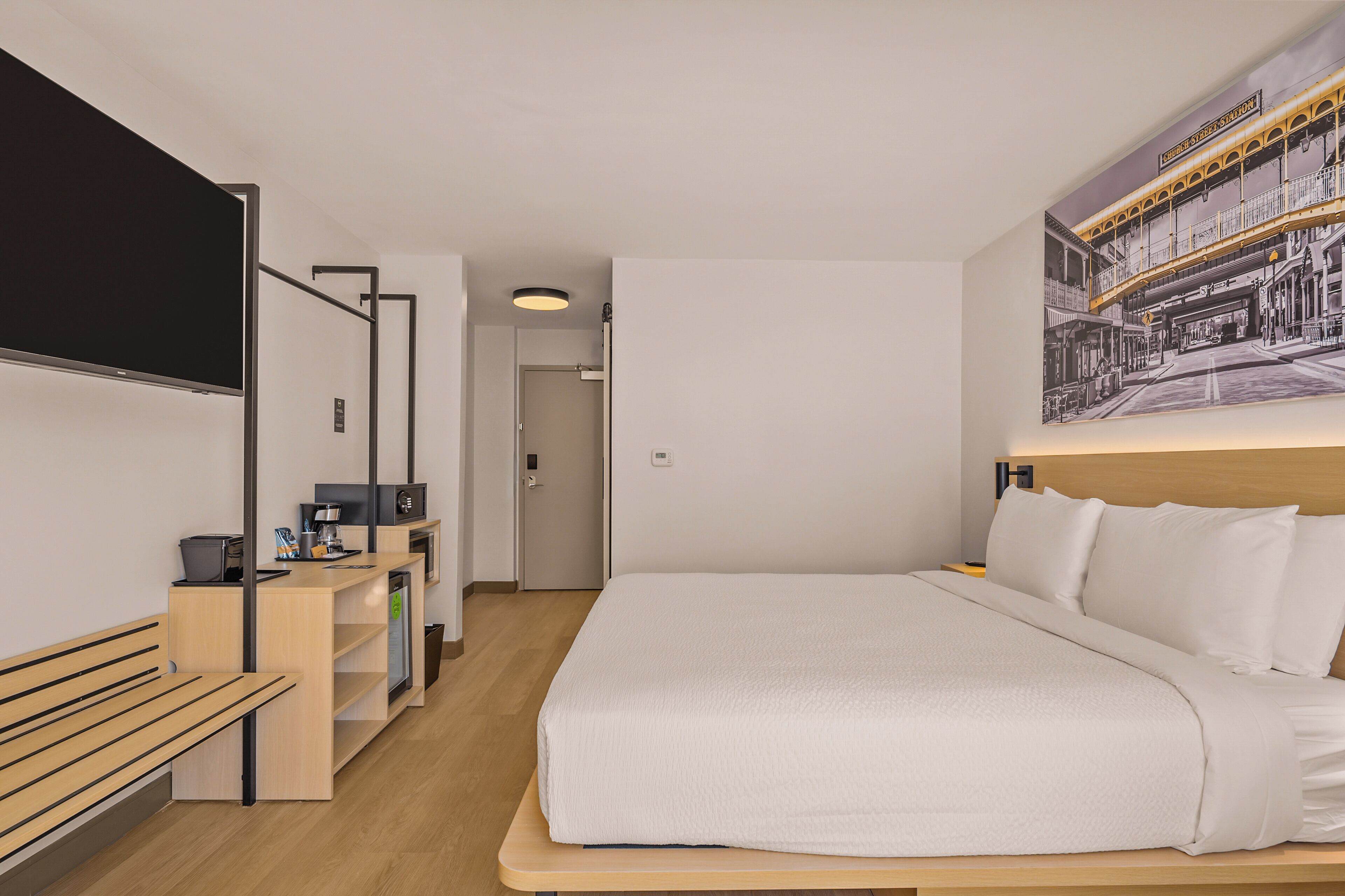 Una habitación de hotel moderna con una cama, un escritorio con computadora, un banco y un televisor montado en la pared.