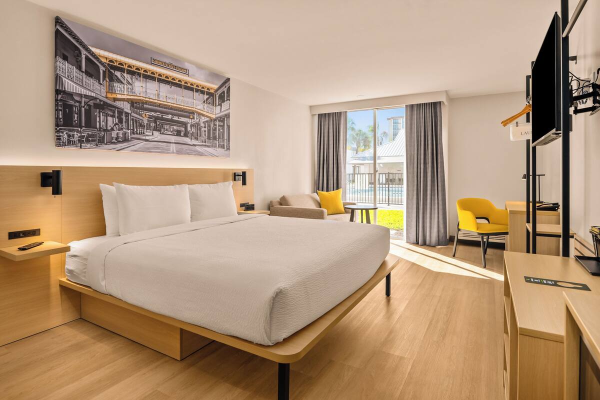 A modern hotel room with a large bed, a desk, a chair, and a balcony with a view of the city.