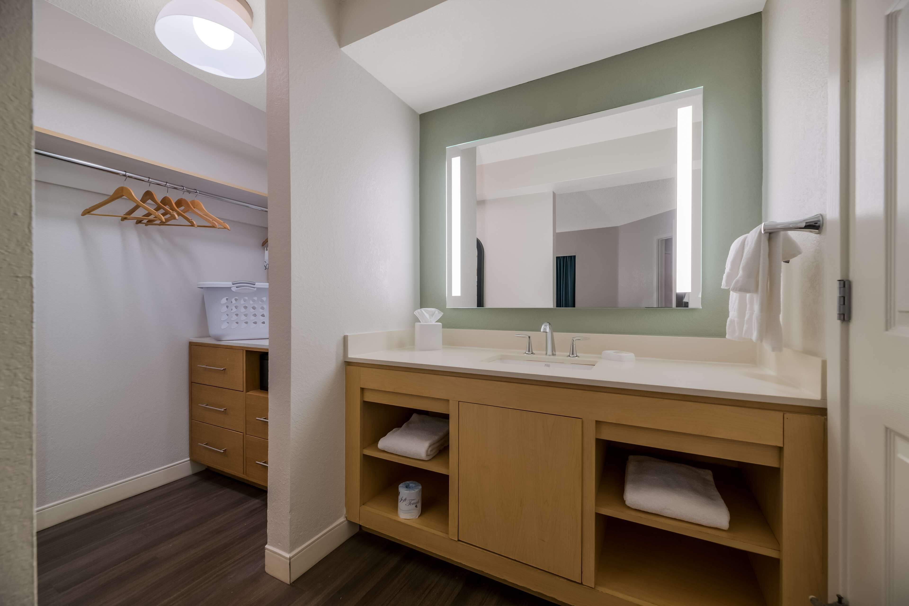 Een moderne badkamer met een grote spiegel, een houten wastafelmeubel en een inloopkast met hangende kleding.