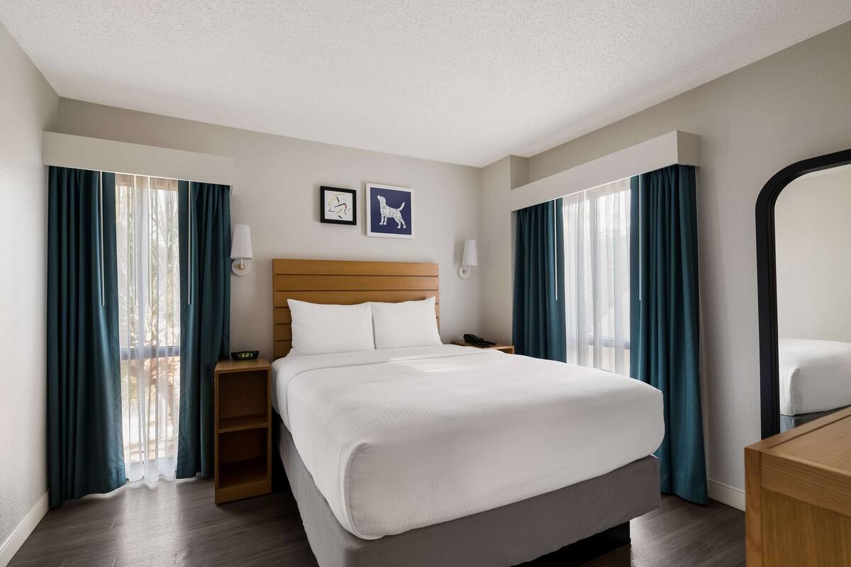 Quarto de hotel com uma cama grande, cabeceira de madeira, duas mesas de cabeceira, um criado-mudo, um espelho e duas fotografias emolduradas na parede.