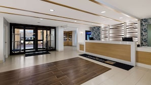 Un espace d’accueil moderne, avec un comptoir de réception en bois, un tapis noir portant le nom « Sanesta », et une vue sur l’espace extérieur grâce à des portes vitrées.