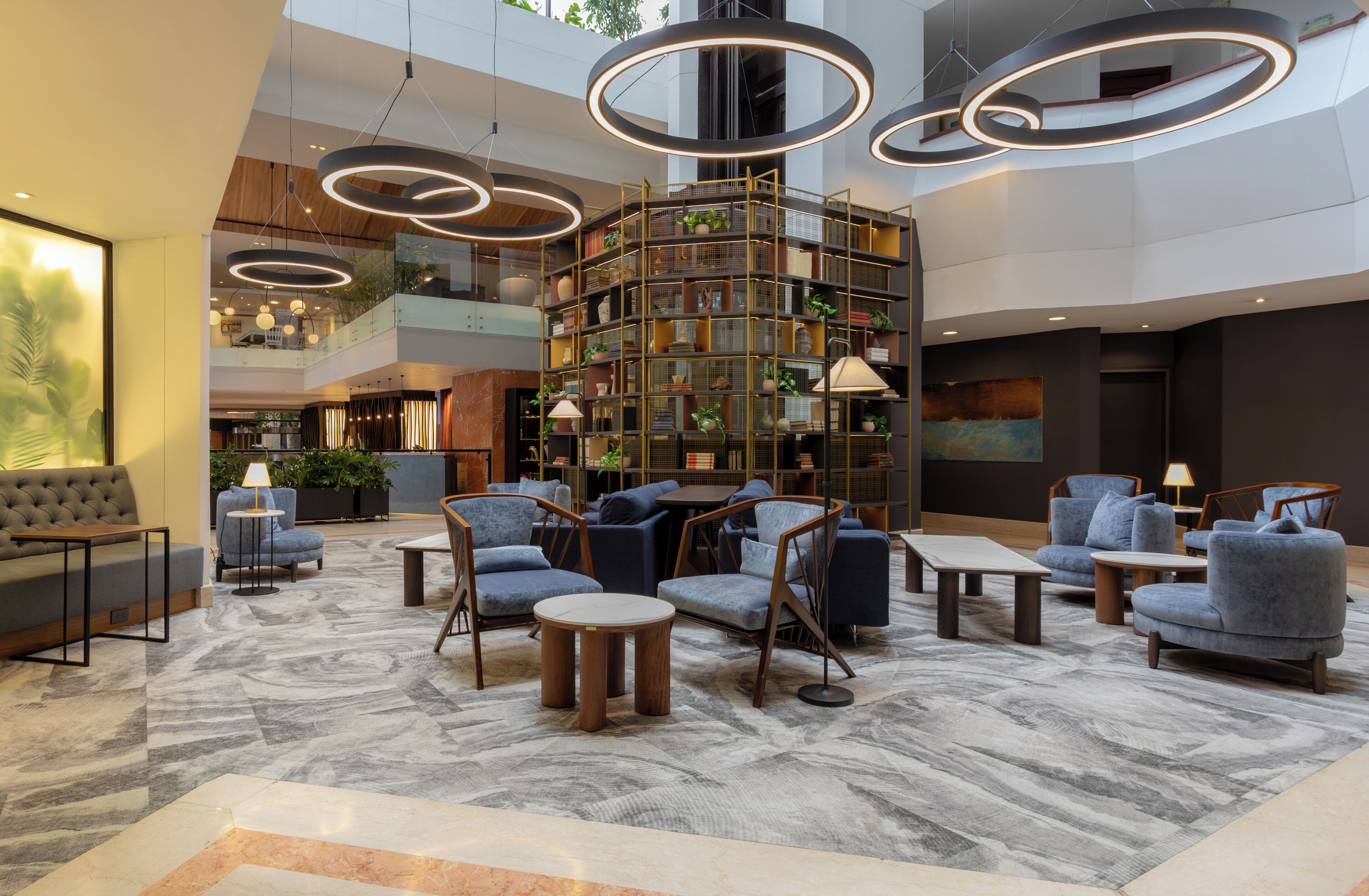 A modern lobby with a central bookshelf, seating area, and a high ceiling with circular light fixtures.