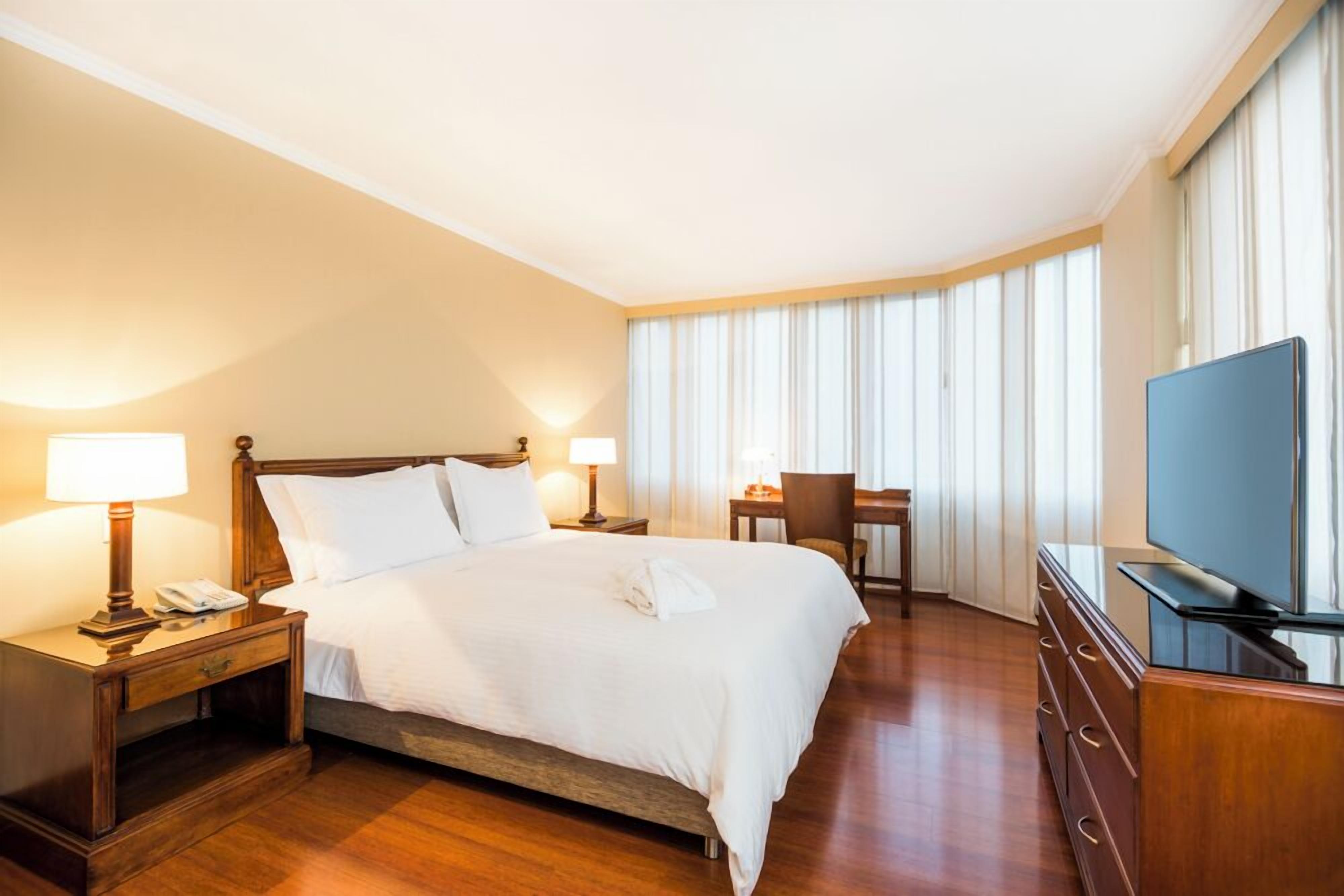 A hotel room with a large bed, bedside tables with lamps, a desk, a chair, and a television.