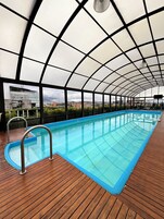 Una piscina techada con cubierta de madera, barandales de metal y vista a edificios al fondo.