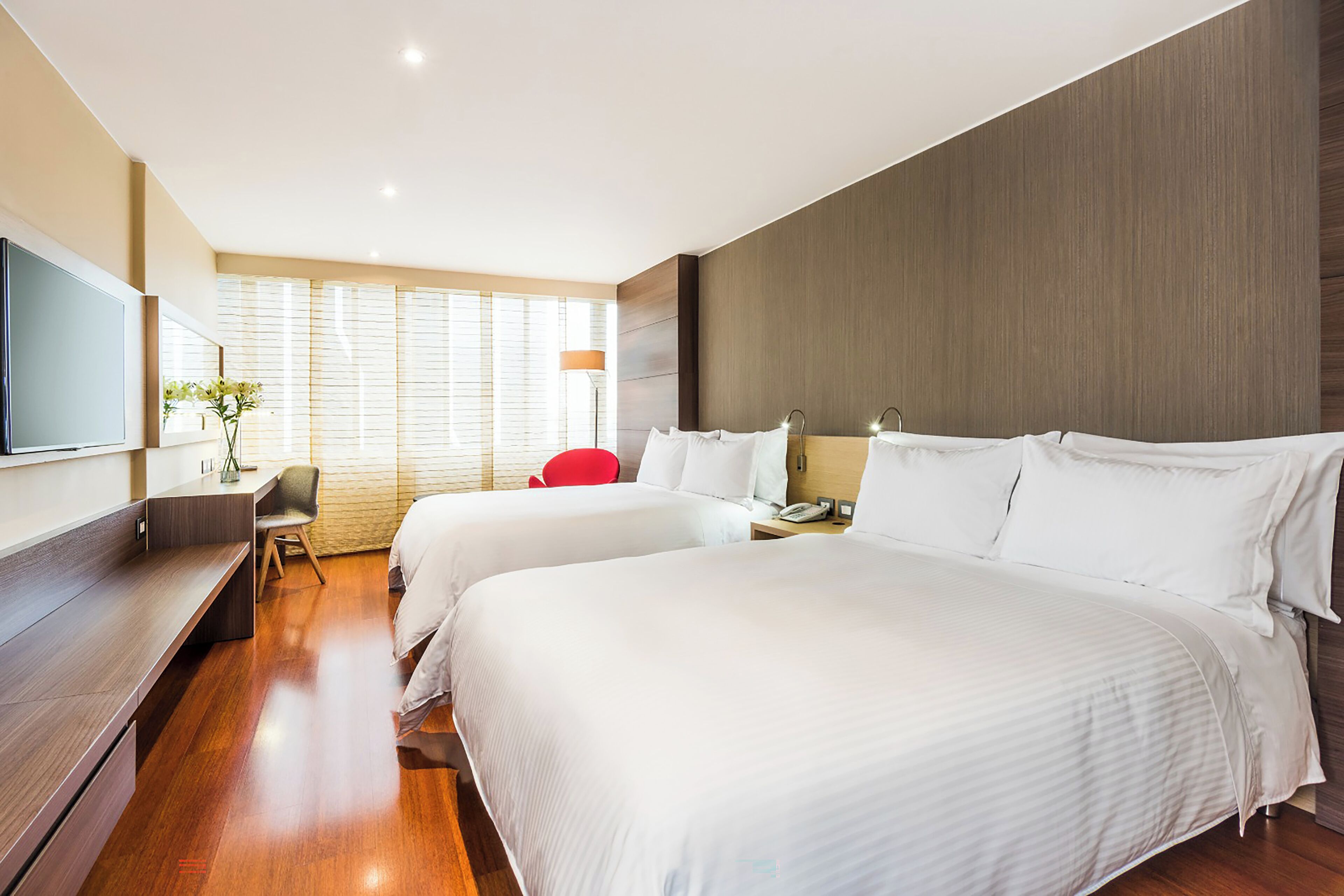 A hotel room with two beds, a desk, a chair, a TV, and a window with blinds.
