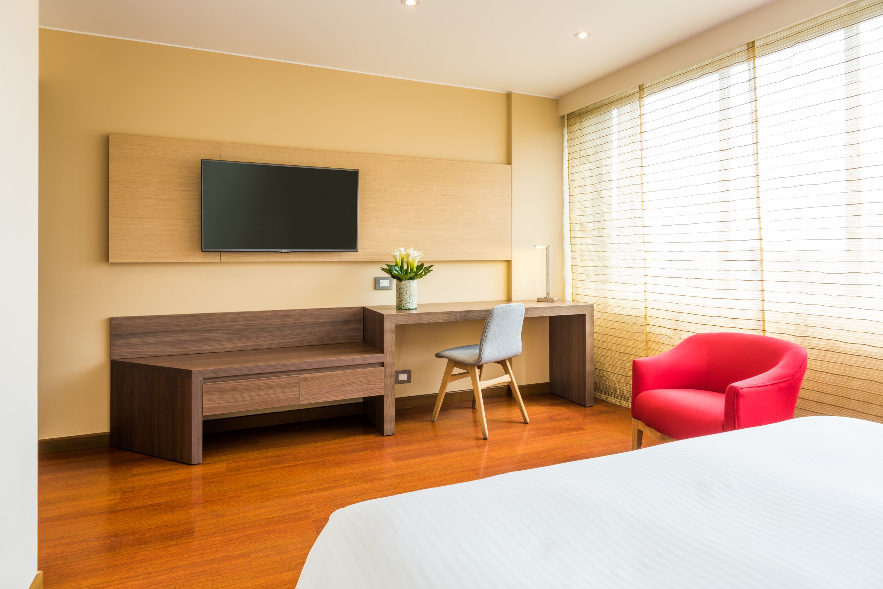Ein modernes Hotelzimmer mit Schreibtisch aus Holz, einem roten Sessel, einem Bett und einem großen Fenster mit Jalousien.