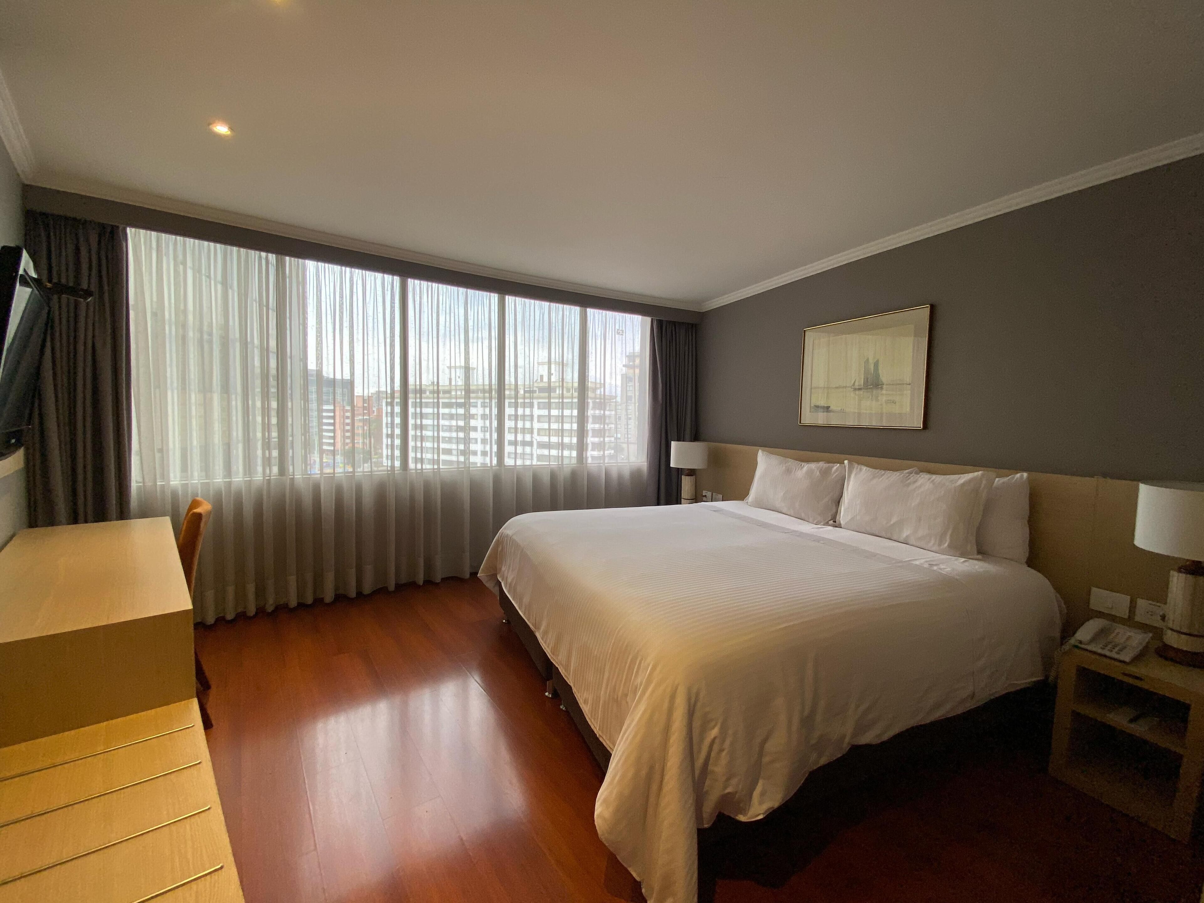 Een hotelkamer met een groot bed, een bureau en uitzicht op de stad door dunne gordijnen.