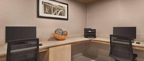 Un espacio de oficina moderno con dos escritorios, cada uno con monitor, teclado y ratón. Hay un escritorio de madera en el centro con un cuenco decorativo encima. La habitación tiene techo de madera y un cuadro en la pared.
