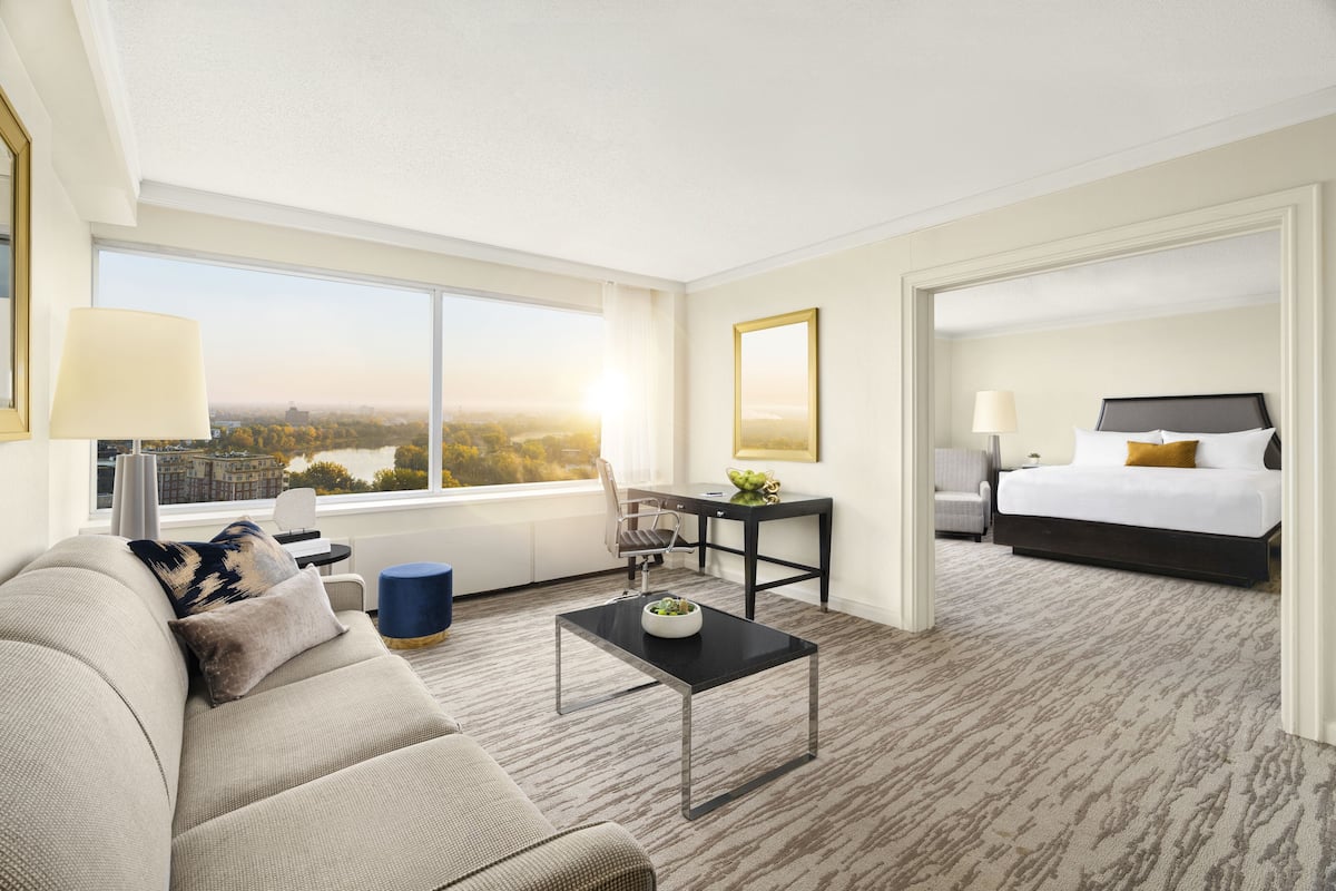 A hotel room with a large window, a sofa, a coffee table, a desk, and a bed.