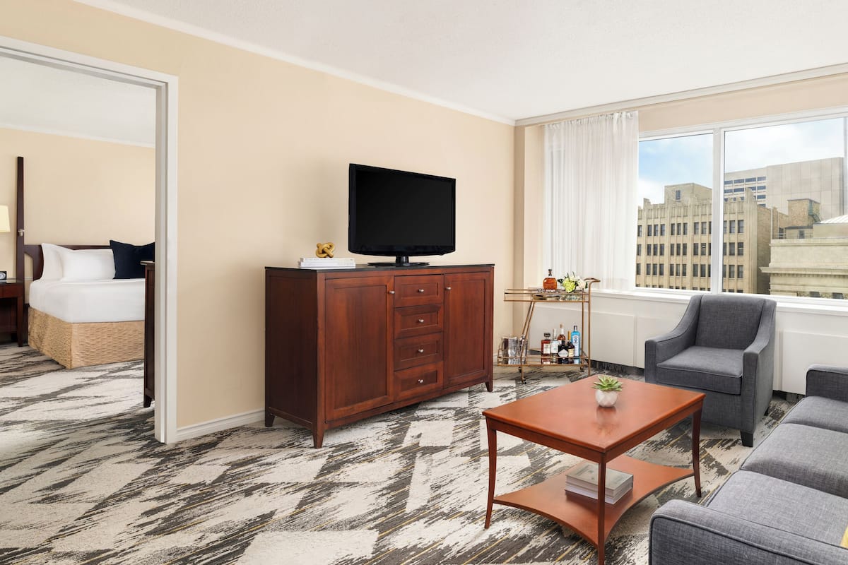 A hotel room with a bed, a television on a wooden cabinet, a grey sofa, a small table, and a window with a city view.