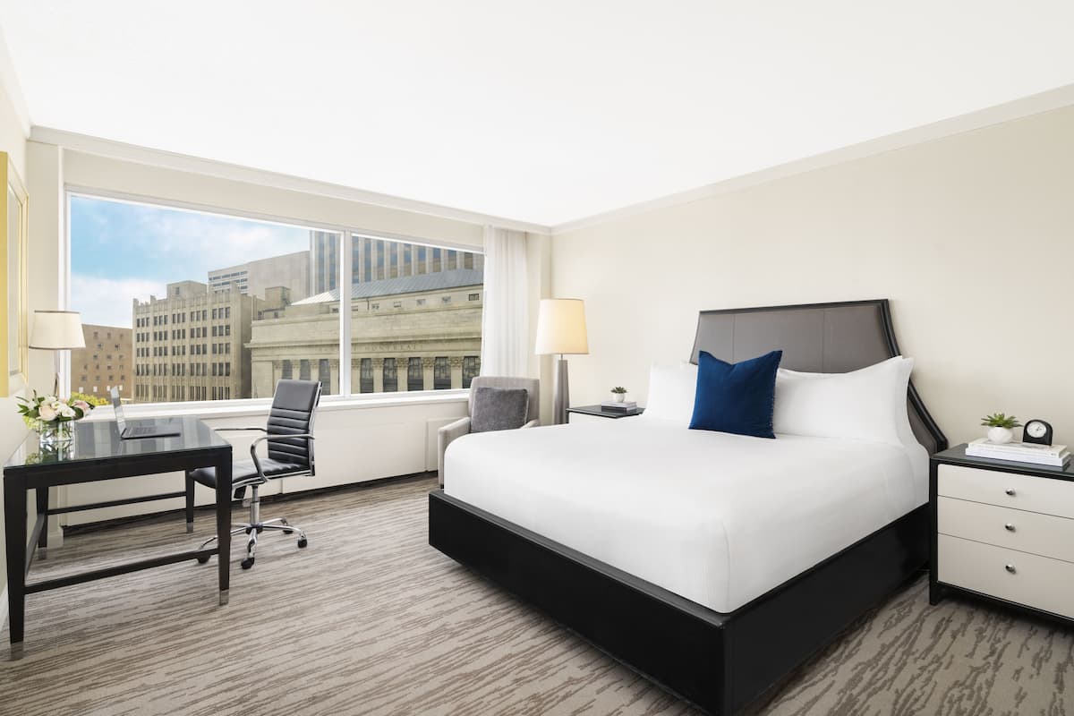A hotel room with a large bed, a desk with a chair, a nightstand, and a window offering a city view.