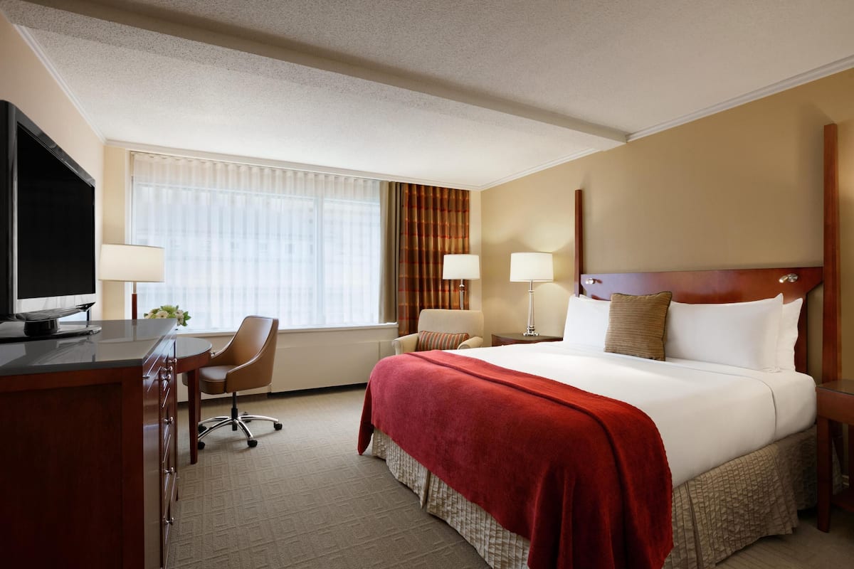 A hotel room with a large bed, a desk with a TV, a chair, and a window with curtains.