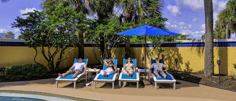 A poolside area with lounge chairs and umbrellas, surrounded by palm trees and a yellow wall.