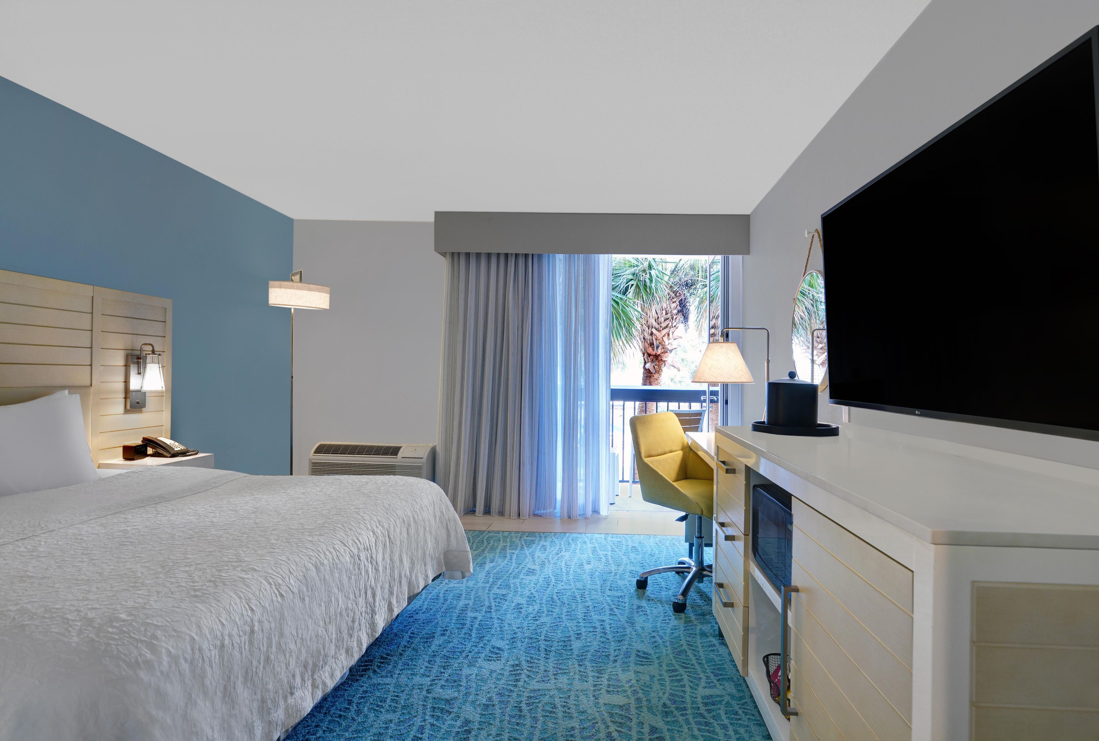 Habitación de hotel con cama, escritorio, silla, televisión y balcón con cortinas.